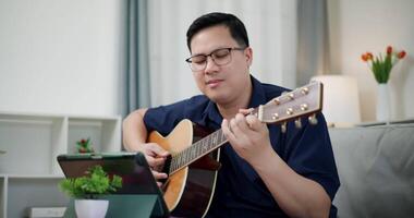 Handheld medium shot, Happy Asian man sitting on sofa enjoying playing music with guitar and looking at music chords on digital tablet in living room with relaxation. video