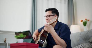Handheld medium shot, Happy Asian man sitting on sofa enjoying playing music with guitar and looking at music chords on digital tablet in living room video