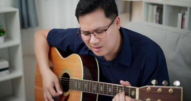 Handheld high angle view shot, An Asian man is sitting comfortably on a sofa, strumming an acoustic guitar and singing happily, enjoying music in a relaxing living room at home. video
