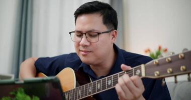 Handheld selective focus shot, Happy Asian man sitting on sofa enjoying playing music with guitar and looking at music chords on digital tablet in living room with relaxation. video