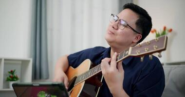 Happy Asian man sit on sofa enjoying playing music with guitar and looking at music chords on digital tablet in living room with relaxation. video