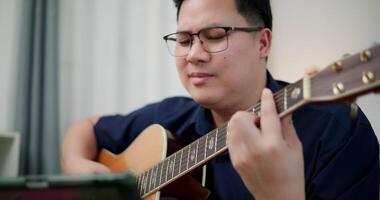 Asian man sitting on sofa enjoy playing music with guitar and looking at music chords on digital tablet in living room with relaxation. video