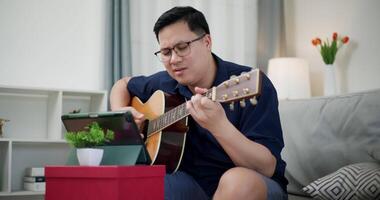 Handheld medium shot, Happy Asian man sitting on sofa enjoying playing music with guitar and looking at music chords on digital tablet in living room at home. video