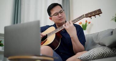 Handheld low angle view shot, Asian man sitting on sofa enjoying playing music with guitar and looking at music chords on laptop in living room video