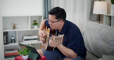 Happy Asian man sit on sofa enjoy play music with guitar and looking at music chords on digital tablet in living room with relaxation. video