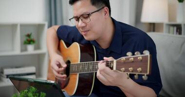 Handheld selective focus shot, Happy Asian man sitting on sofa enjoying playing music with guitar and looking at music chords on digital tablet in living room at home video