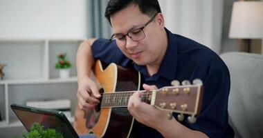 Happy Asian man sit on sofa enjoy playing music with guitar and looking at music chords on digital tablet in living room with relaxation. video