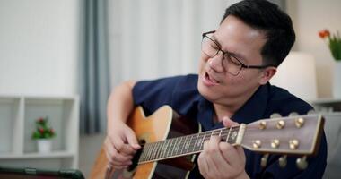 Handheld shot, Happy Asian man sitting on sofa enjoying playing music with guitar and looking at music chords on digital tablet in living room with relaxation. video
