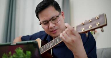 Handheld selective focus shot, Happy Asian man sitting on sofa enjoying playing music with guitar and looking at music chords on digital tablet in living room video
