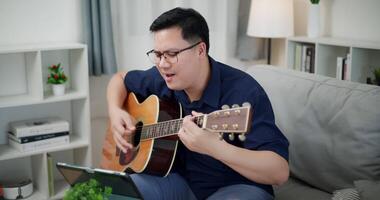 Handheld shot, Happy Asian man sit on sofa enjoying playing music with guitar and looking at music chords on digital tablet in living room with relaxation. video