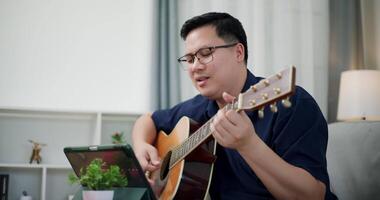 Handheld shot, Happy man sitting on sofa enjoying playing music with guitar and looking at music chords on digital tablet in living room with relaxation. video