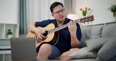 Handheld medium shot, Asian man sitting on sofa enjoying playing music with guitar and looking at music chords on laptop in living room with relaxation. video