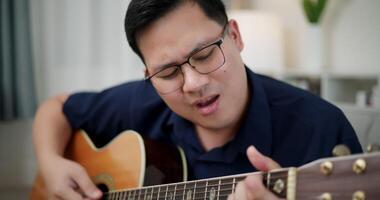 Handheld shot, Asian man sitting on sofa enjoying playing music with guitar and looking at music chords on digital tablet in living room with relaxation. video