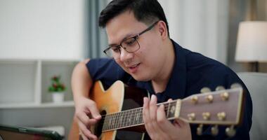 Handheld selective focus shot, Asian man sitting on sofa enjoying playing music with guitar and looking at music chords on digital tablet in living room with relaxation. video