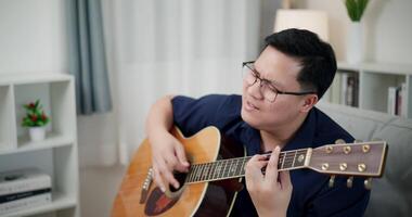 Happy Asian man sitting on sofa enjoying playing music with guitar and looking at music chords on digital tablet in living room with relaxation. video