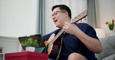 Handheld low angle view shot, Happy Asian man sitting on sofa enjoying playing music with guitar and looking at music chords on digital tablet in living room with relaxation. video