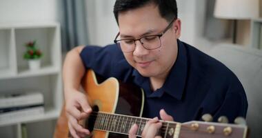 Handheld selective focus shot,Happy Asian man sitting on sofa enjoying playing music with guitar and looking at music chords on digital tablet in living room with relaxation. video