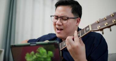 Handheld shot,Happy Asian man sitting on sofa enjoying playing music with guitar and looking at music chords on digital tablet in living room with relaxation. video
