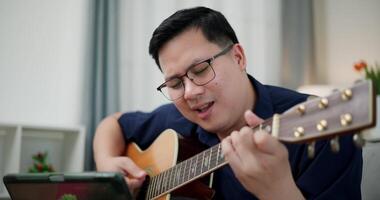Asian man sitting on sofa enjoying playing music with guitar and looking at music chords on digital tablet in living room with relaxation. video