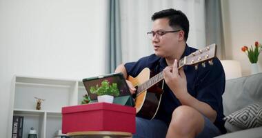 Handheld low angle view shot,Happy Asian man sitting on sofa enjoying playing music with guitar and looking at music chords on digital tablet in living room with relaxation. video