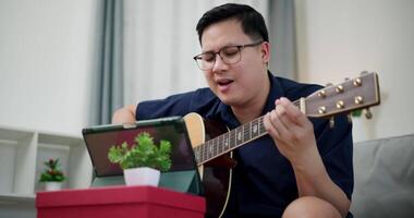 Handheld medium shot,Happy Asian man sitting on sofa enjoying playing music with guitar and looking at music chords on digital tablet in living room with relaxation. video
