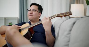 Handheld low angle view shot, An Asian man is sitting on a sofa, strumming a guitar and singing happily, enjoying music in a comfortable living room at home. video