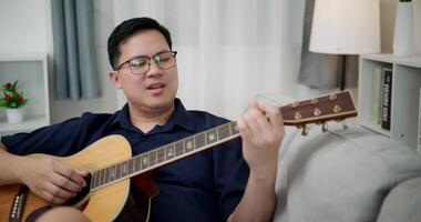Handheld shot, An Asian man is sitting on a sofa, strumming a guitar and singing happily, enjoying music in a comfortable living room at home. video