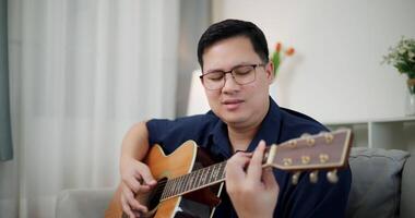 Handheld shot,An Asian man is sitting on a sofa, strumming a guitar and singing happily, enjoying music in a comfortable living room at home. video