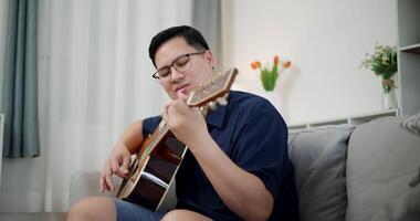 Handheld shot, Relaxed Asian man wearing glasses and a navy-blue shirt is enjoy playing an acoustic guitar on a sofa at home. video