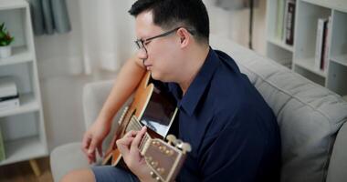 Handheld high angle view shot, Relaxed Asian man wearing glasses and a navy-blue shirt is enjoy playing an acoustic guitar on a sofa at home. video