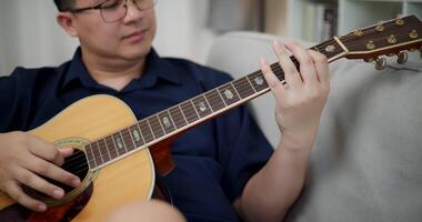 Handheld selective focus shot, Relaxed Asian man happily playing music with guitar on sofa at home. video