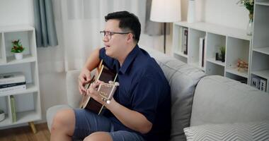Relaxed Asian man happily playing music with guitar on sofa at home. video