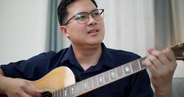 Handheld selective focus shot, An Asian man is sitting on a sofa, strumming a guitar and singing happily, enjoying music in a comfortable living room. video