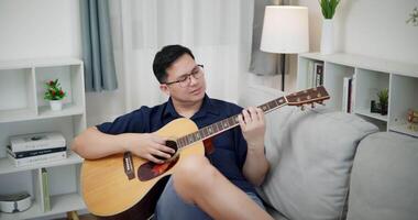 Handheld high angle view shot, An Asian man is sitting on a sofa, strumming a guitar and singing happily, enjoying music in a comfortable living room video