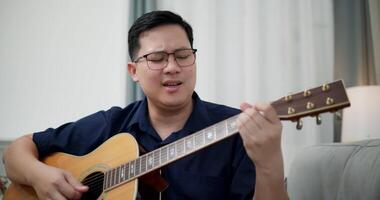 Handheld shot, An Asian man is sitting on a sofa, strumming a guitar and singing happily, enjoying music in a comfortable living room. video