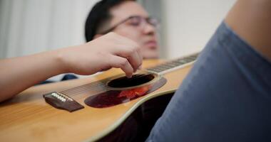 Handheld selective focus shot, An Asian man is sitting on a sofa, strumming a guitar and singing happily, enjoying music in a comfortable living room at home. video