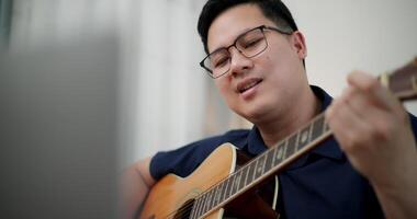Asian man sit on sofa enjoying playing music with guitar and looking at music chords on laptop in living room with relaxation. video