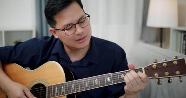 Asian man is sitting on a sofa, strumming a guitar and singing happily, enjoying music in a comfortable living room at home. video