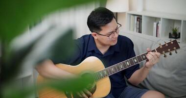 Handheld high angle view shot, Asian man is sitting on a sofa, strumming a guitar and singing happily, enjoying music in a comfortable living room at home. video