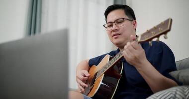 Handheld low angle view shot, Asian man sitting on sofa enjoying playing music with guitar and looking at music chords on laptop in living room with relaxation. video