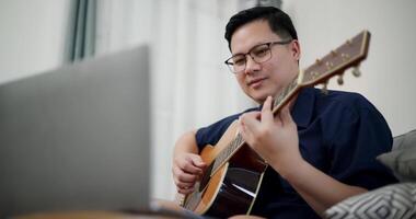 Handheld low angle view shot, Relaxed Asian man sitting on sofa enjoying playing music with guitar and looking at music chords on laptop in living room with relaxation. video