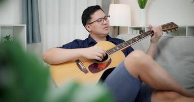 Handheld selective focus shot, An Asian man is sitting on a sofa, strumming a guitar and singing happily, enjoying music in a cozy living room at home. video