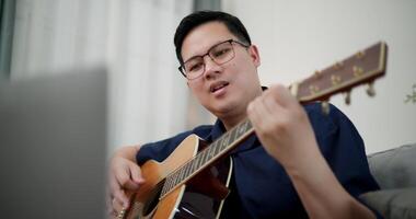 Handheld shot, Asian man sitting on sofa enjoying playing music with guitar and looking at music chords on laptop in living room with relaxation. video