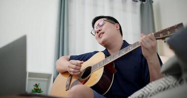 Handheld low angle view shot,Asian man sitting on sofa enjoying playing music with guitar and looking at music chords on laptop in living room with relaxation. video