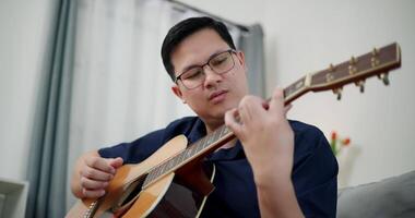 Handheld low angle view shot, Relaxed Asian man wearing glasses and a navy-blue shirt is enjoy playing an acoustic guitar on a sofa in the living room at home. video