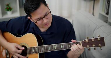 Handheld high angle view shot, Happy Asian man is sitting on a sofa, strumming a guitar and singing happily, enjoying music in a comfortable living room at home. video