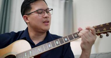 Handheld selective focus shot, Asian man is sitting on a sofa, strumming a guitar and singing happily, enjoying music in a comfortable living room at home. video