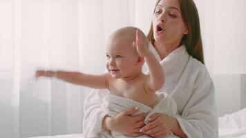 A mother lovingly wraps her toddler in a soft towel while sitting on a comfortable surface at home. The warm light creates a cozy atmosphere, highlighting their connection and joy. video