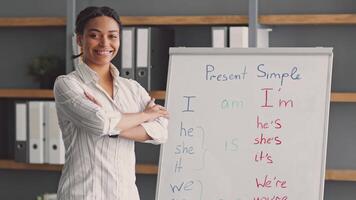 An African American woman stands confidently by a whiteboard, teaching a remote class about the present simple tense. She uses clear examples to help her students understand. video