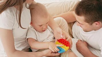 A young child sits on a bed, focused on a colorful toy as both parents assist with playtime. The comfortable indoor setting highlights family bonding during a learning activity. video